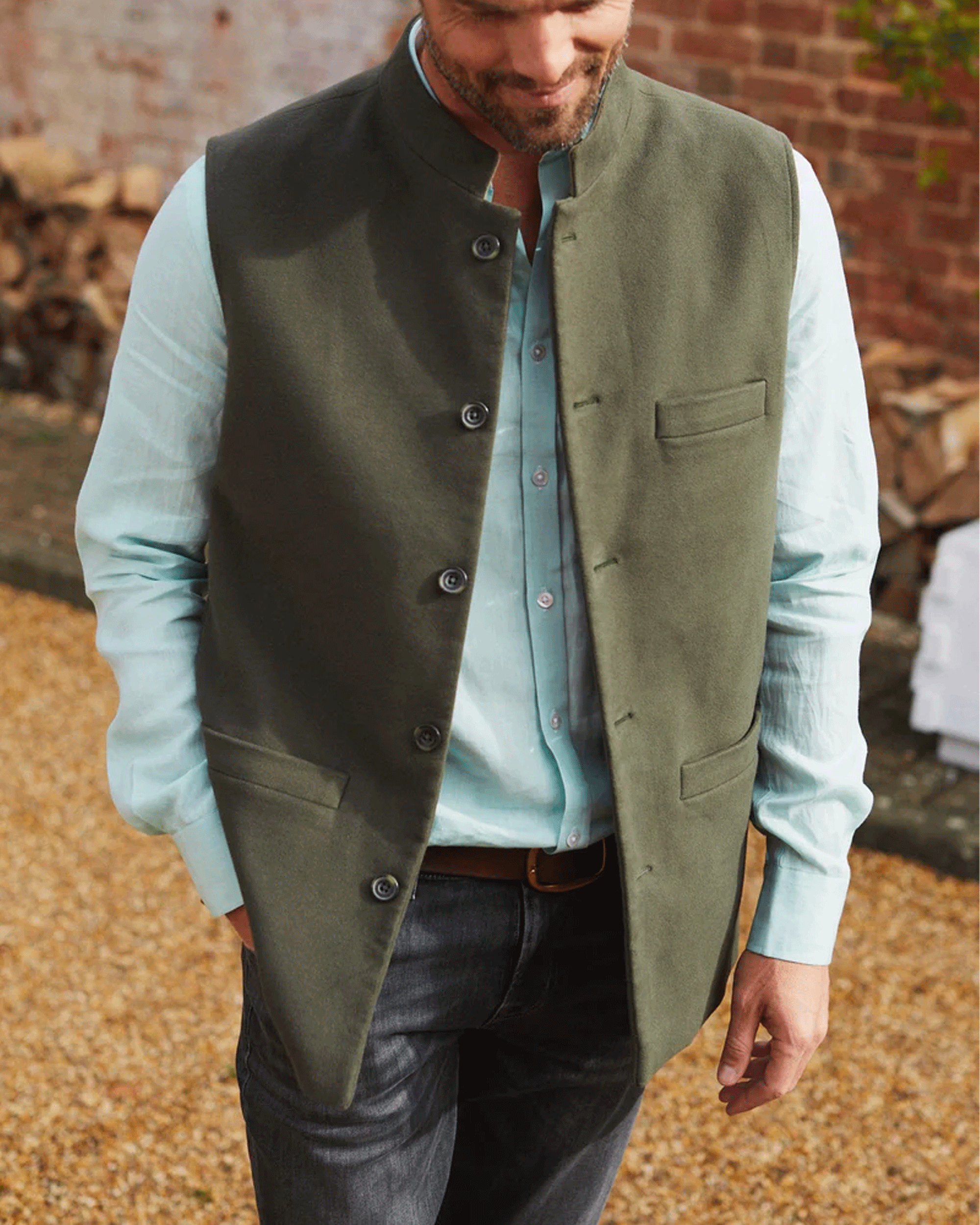 A man wearing an olive green waistcoat vest over a light blue button-down shirt and dark gray jeans, standing outdoors on a gravel path with a brick wall in the background.