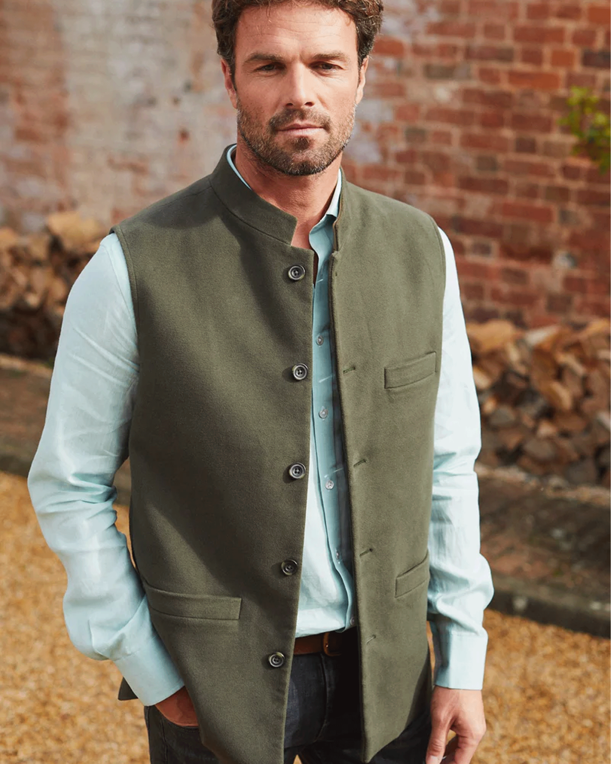 A man wearing an olive green waistcoat over a light blue dress shirt, standing outdoors in front of a brick wall.