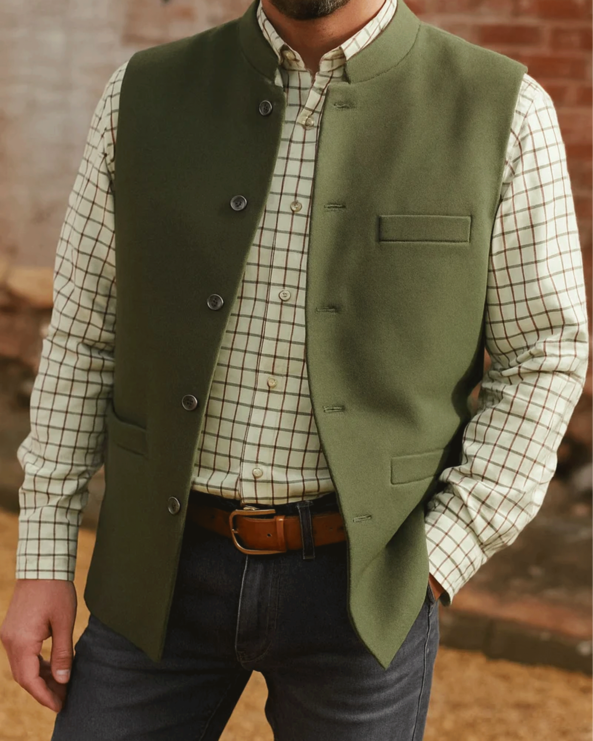 A neck to thigh photo of a man wearing a olive green nehru ilet over a checked shirt with blue jeans and a brown belt