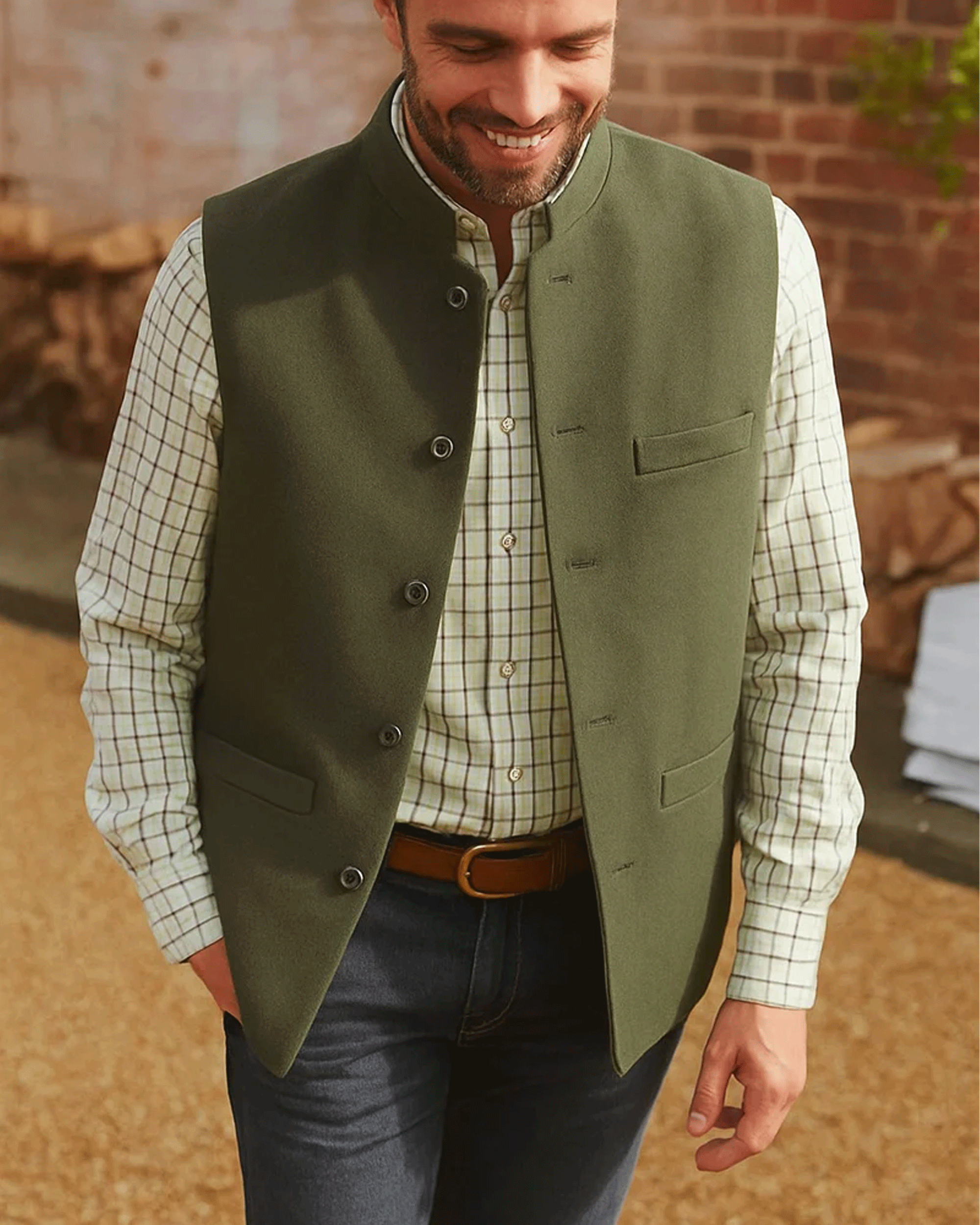 A man wearing an olive green Nehru jacket vest over a cream and black checkered shirt, paired with dark blue jeans and a brown belt, walking outdoors against a brick wall background.