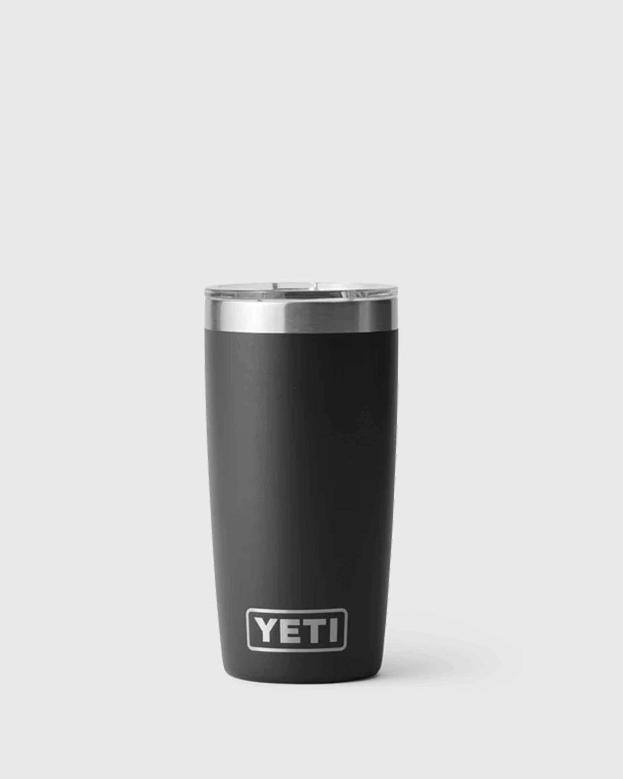A black insulated tumbler with a clear lid and stainless steel rim, standing upright against a light grey background. The tumbler features the YETI logo near the bottom and has a tapered shape that narrows toward the base.