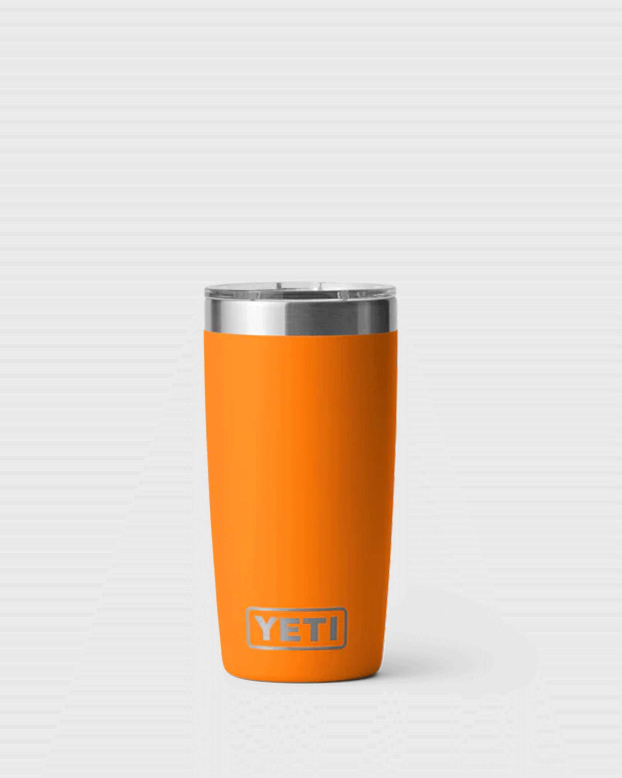 A bright orange insulated YETI tumbler called King Crab with a stainless steel rim and clear plastic lid, standing upright against a light grey background. The tumbler has a smooth matte finish, a slightly tapered shape, and the YETI logo near the bottom front.