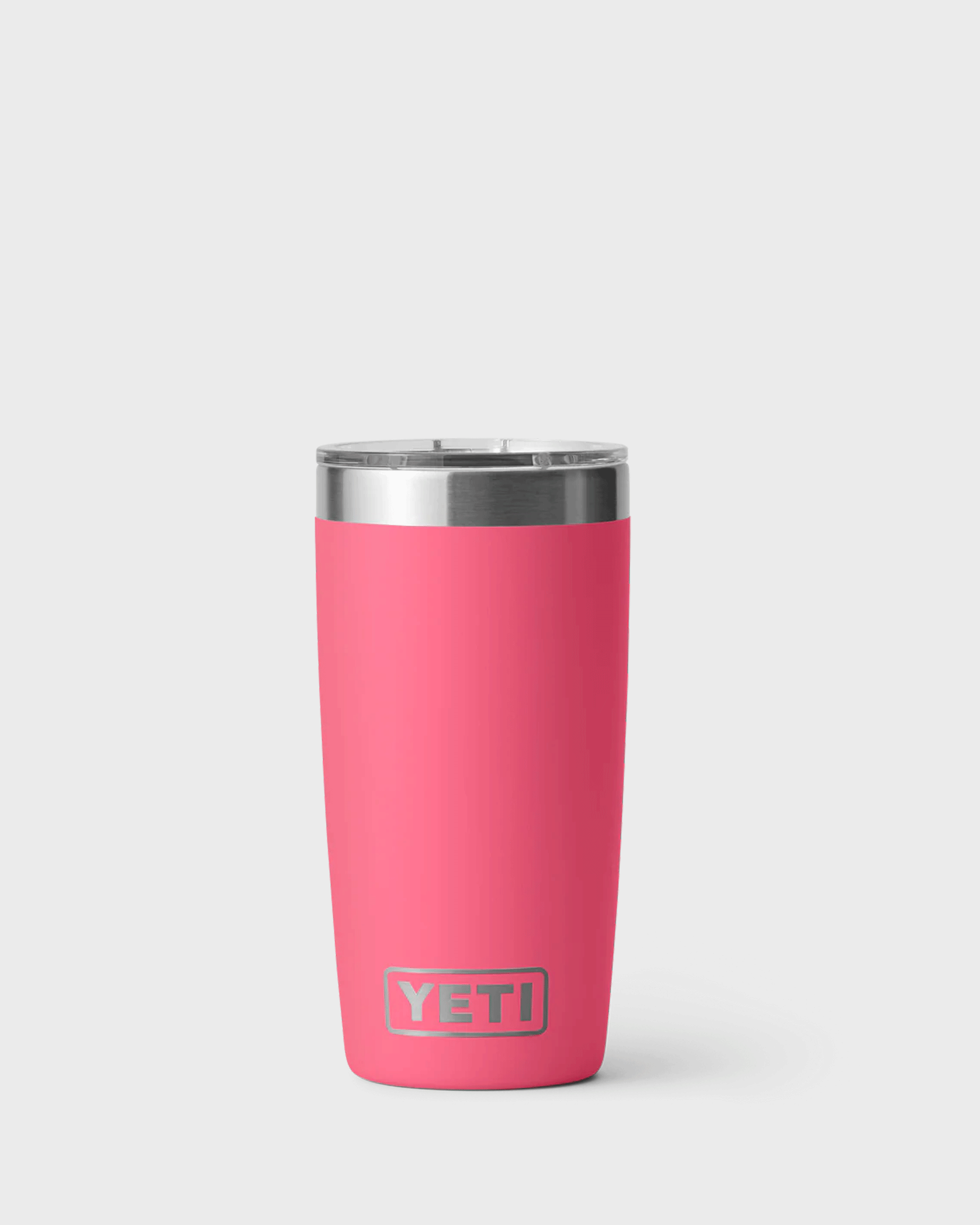 A vibrant bright pink insulated YETI tumbler called Tropical Pink has a stainless steel rim and clear plastic lid, standing upright against a light grey background. The tumbler has a smooth matte finish, a slightly tapered shape, and the YETI logo near the bottom front.