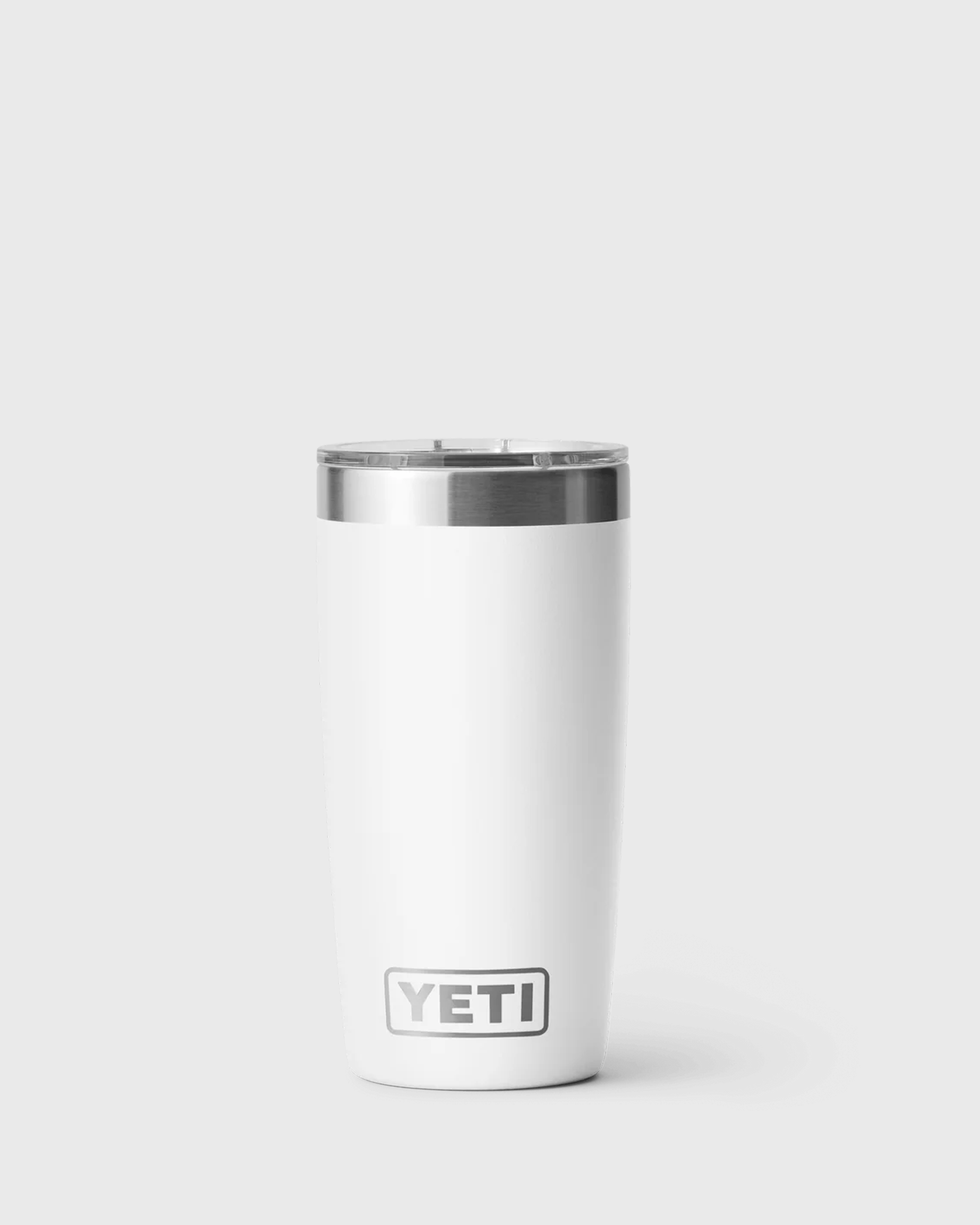 A white insulated tumbler with a clear lid and stainless steel rim, standing upright against a light grey background. The tumbler features the YETI logo near the bottom and has a tapered shape that narrows toward the base.