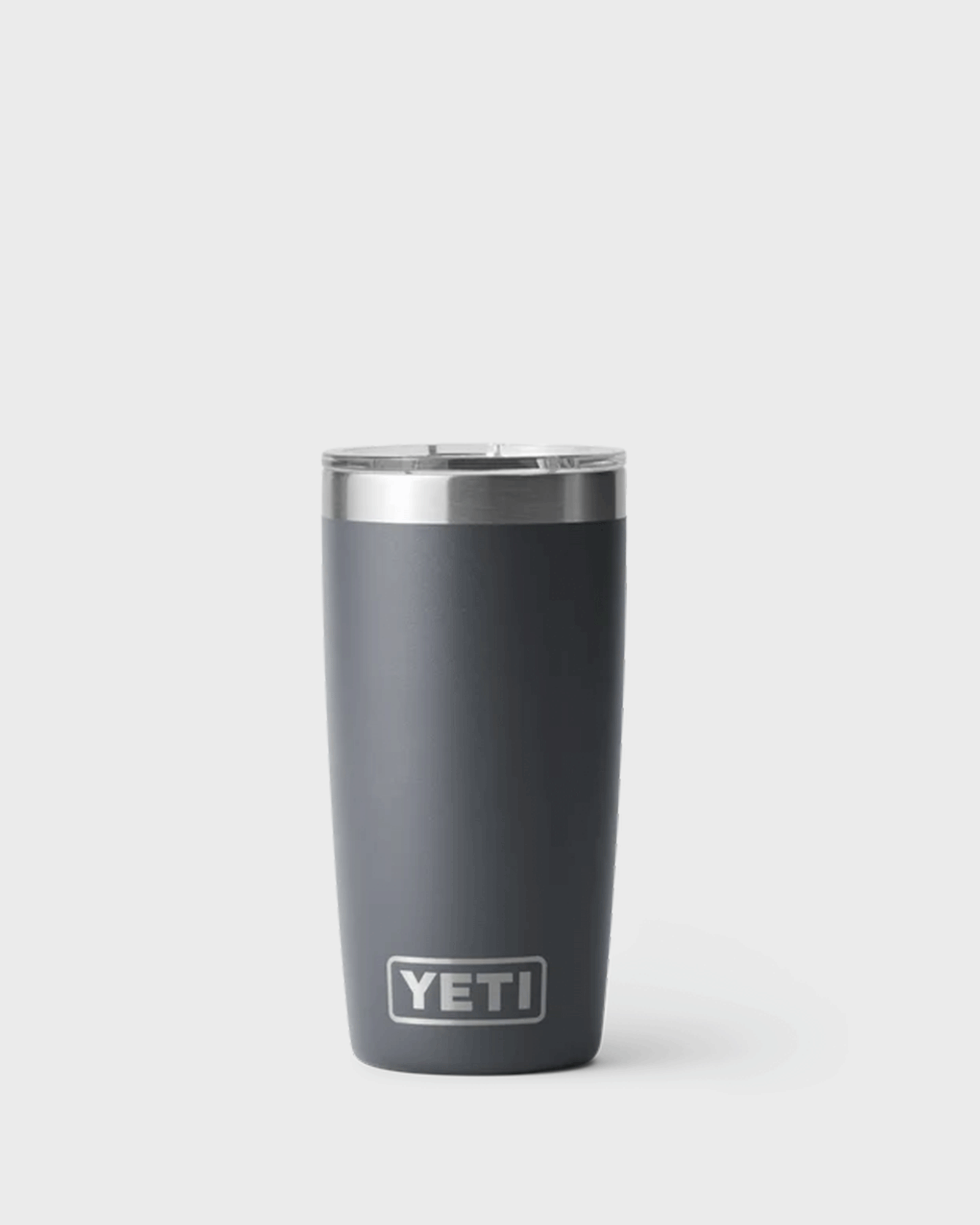 A charcoal insulated YETI tumbler with a stainless steel rim and clear plastic lid, standing upright against a light grey background. The tumbler has a smooth matte finish, a slightly tapered shape, and the YETI logo near the bottom front.