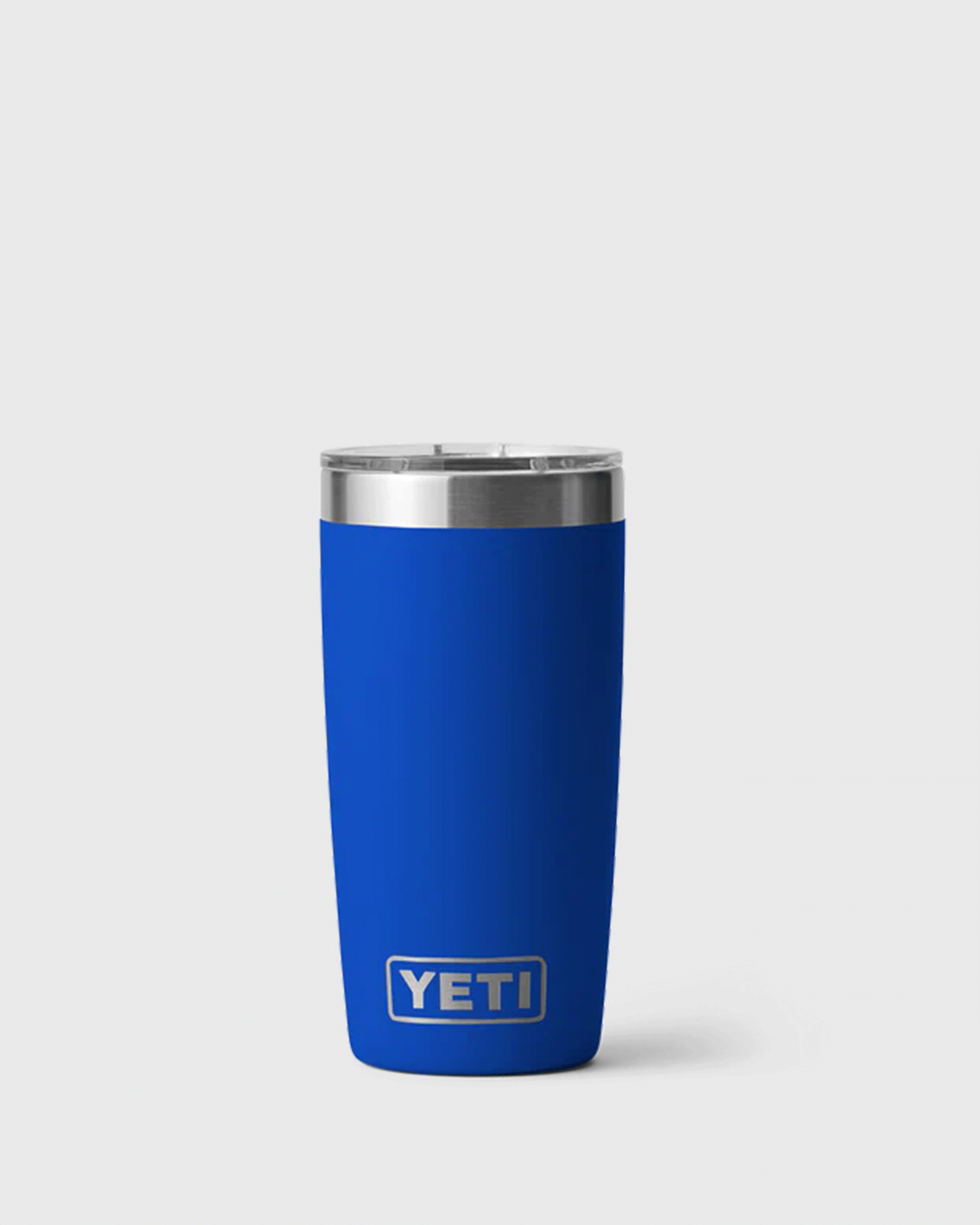A bright blue insulated tumbler with a clear lid and stainless steel rim, standing upright against a light grey background. The Royal Blue tumbler features the YETI logo near the bottom and has a tapered shape that narrows toward the base.