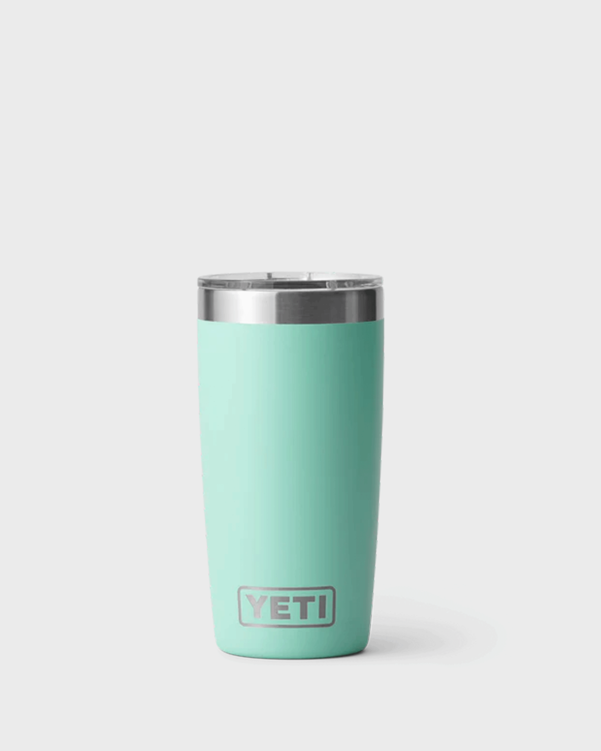A mint green insulated YETI tumbler called Seafoam with a stainless steel rim and clear plastic lid, standing upright against a light grey background. The tumbler has a smooth matte finish, a slightly tapered shape, and the YETI logo near the bottom front.