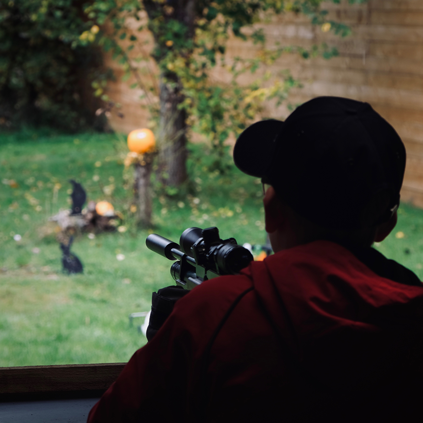 A person with a red jacket and black hat is seated, aiming an air rifle at a target in the distance.
